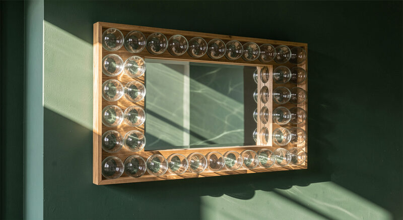 A rectangular mirror framed with rows of clear glass domes on a dark green wall, side light raking across the domes.