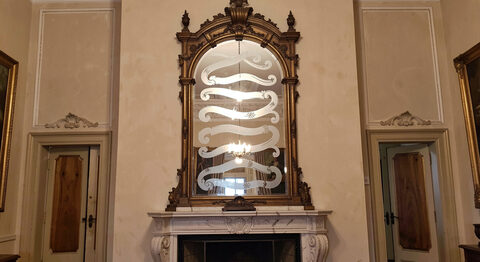 A tall crowned Venetian mirror hanging above a marble fireplace, etched glass scrolls catching the light