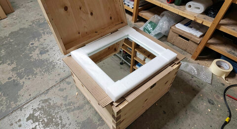 An open wooden crate with a mirror wrapped in foam and corrugated board, packing tape and bubble wrap on the floor