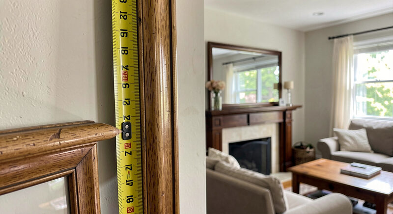 A measuring tape held against a wall beside a rectangular mirror, a pencil mark at the height of a mantel in a living room.