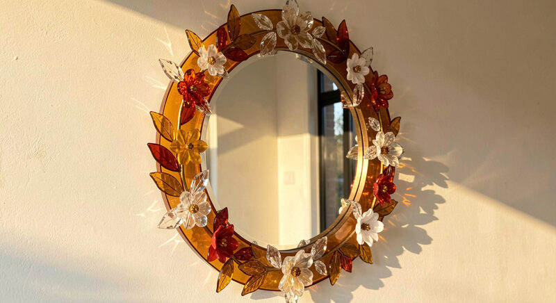 A round mirror framed with amber and crystal glass leaves and applied flowers on a pale wall in warm afternoon light.