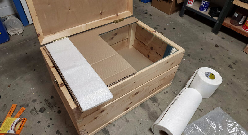 An open wooden crate on a workshop floor with a mirror wrapped in foam and corrugated board, packing tape and a roll of bubble wrap nearby.