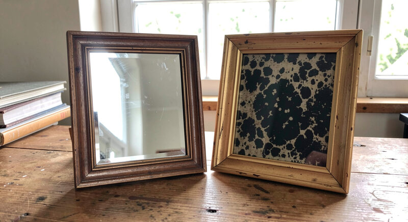 Two small framed mirrors side by side on a wooden table, one bright and clear and one with a dark mottled antique surface, daylight from a window.