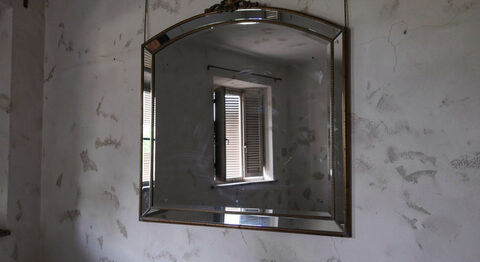 A large antique Venetian mirror with a bevelled glass frame hanging on a bare plaster wall in a dim room, a single shuttered window reflected in the glass.