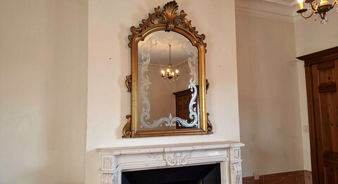 A tall crowned Venetian mirror above a fireplace in a period room, its etched glass scrolls catching the light of a small chandelier.