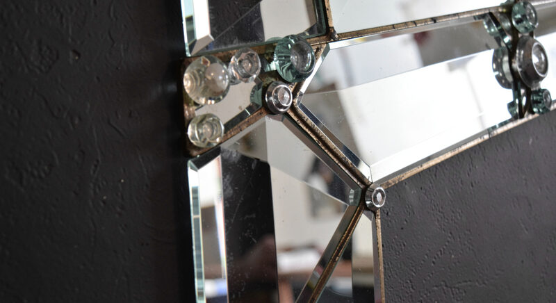 A close view of the corner of a Venetian mirror showing bevelled glass pieces joined with small glass rosettes over a dark wall.